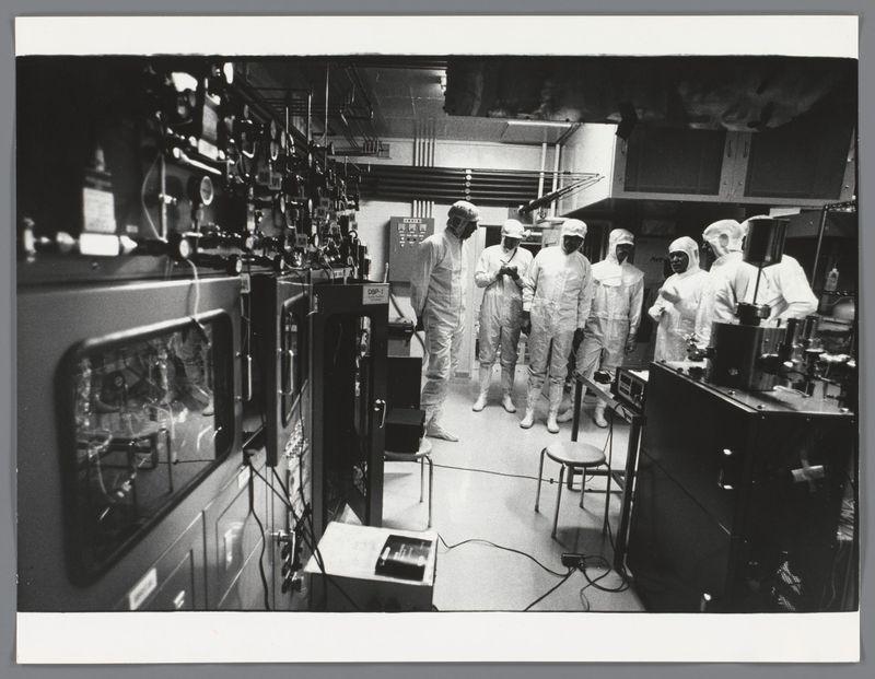 This image shows a group of people dressed in white protective suits and headgear, working together inside what appears to be a laboratory or industrial setting. The environment is filled with various machines and equipment, suggesting that they are engaged in some form of scientific research or technical work. Some individuals appear focused on their tasks while others seem to be observing the activities around them. The black-and-white photograph conveys an atmosphere reminiscent of mid-20th-century technological advancements.

Additional information about this image can be found at Excursie met Groningse studenten en mannen van een organisatie in Japan, in pakken in een laboratorium, Ed van der Elsken, 1985 - 1990 https://images.loener.nl/EdvdElsken/full/6484/64844a0cba25398d908336f4.jpg. This indicates that the photo was taken during a trip involving students and men from an organization in Japan, captured by Ed van der Elsken between 1985-1990.