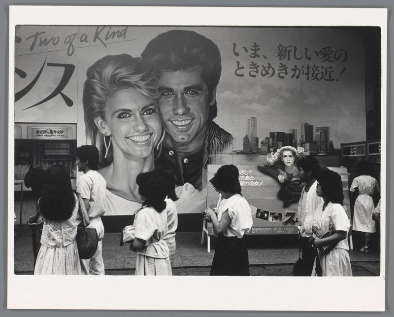 The image is a black and white photograph capturing a moment in an urban setting. It features several people, primarily women dressed in light-colored dresses or skirts with dark tops, standing on a sidewalk. They appear to be engaged in various activities: some are looking at something out of frame, others seem to be conversing quietly.

In the background is a large advertisement poster for "Grease," displaying actors Olivia Newton-John and John Travolta's faces along with Japanese text that suggests it translates roughly as "Two of a Kind" or "A Pair." The backdrop shows what appears to be part of an office building, adding context to the urban environment. There are also hints of other posters in the vicinity.

The image evokes a sense of daily life and leisurely activities amidst a bustling cityscape with popular culture elements permeating public spaces.