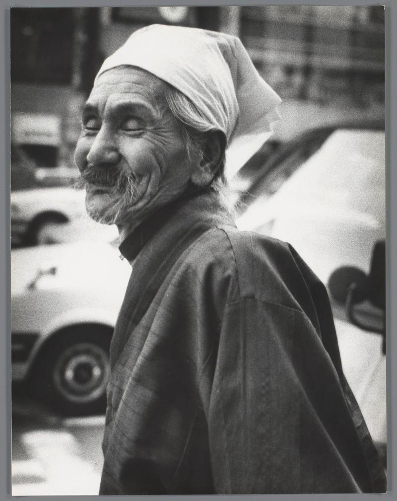 The image depicts an elderly man with a joyful expression, his eyes half-closed and mouth slightly open as if he's laughing or smiling. He has a mustache and is wearing what appears to be traditional clothing that includes a headscarf draped over one shoulder. The background shows urban scenery featuring vehicles, indicating the photo was taken on a street in an urban area. Given this description and additional context provided by the link (Portret van een onbekende oude man op straat, Japan), it is likely that the photograph captures a candid moment of joy or amusement captured by Dutch photographer Ed van der Elsken during his travels.