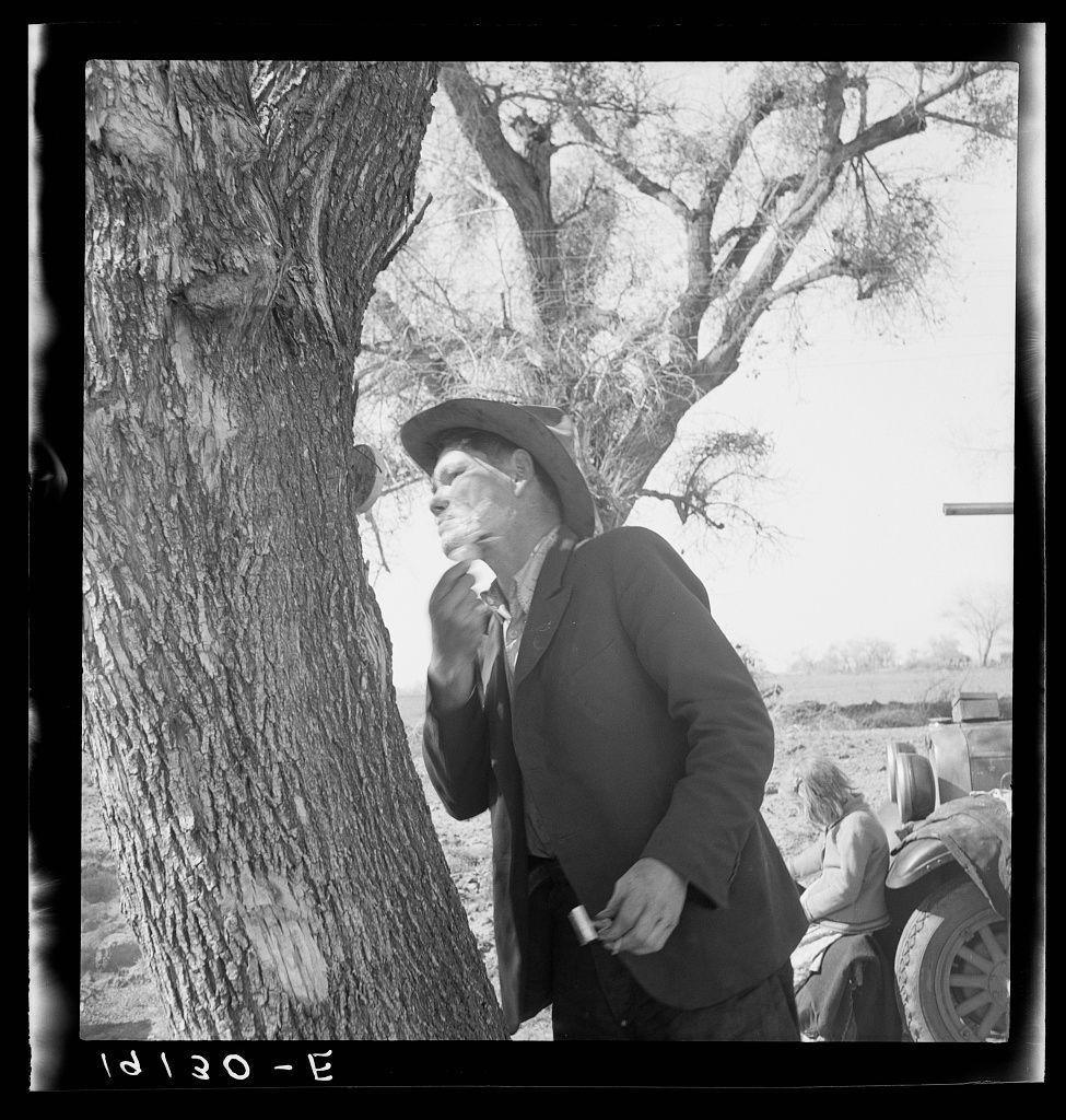 The image captures a black and white photograph of an older man standing by the roadside, engaged in shaving with a manual razor. He is wearing glasses and dressed in what appears to be work clothing suitable for outdoor labor or farming activities. The background shows sparse vegetation and a partly cloudy sky, suggesting an arid landscape typical of rural regions during migration periods.

A tractor trailer can be seen parked behind him, indicating that he might have been traveling with others who are also working as migrant farmworkers. Another individual is visible in the far distance next to the vehicle, which adds context about the transient nature of their situation. The date "1930 - E" suggests this photograph was taken during a significant period for labor movements and rural life conditions.

The overall scene portrays an intimate moment of daily routine amidst challenging circumstances faced by migrant farmworkers in early 20th-century America, reflecting Dorothea Lange's commitment to documenting the harsh realities of working-class lives.
