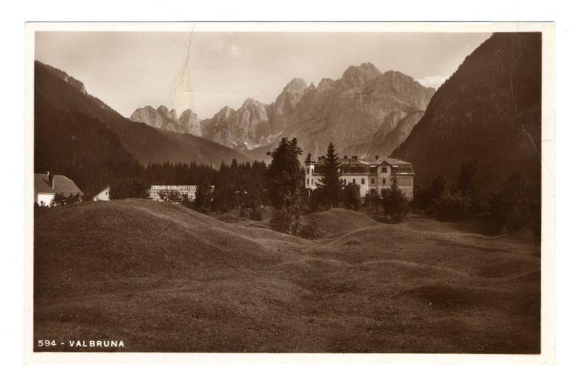 The image is a sepia-toned photograph depicting a panoramic view of Valbruna, with its prominent mountain range in the background. The mountains rise majestically against an overcast sky, and their rocky peaks are highlighted by what appears to be sunlight or possibly distant clouds. In the foreground, gentle undulations of grassy hills create a textured landscape leading up to a cluster of buildings nestled among trees.

The architectural style of these structures suggests historical significance, likely European in origin given its context within Valbruna. The central building is large and has multiple gables with what seem like windows or glass panes reflecting the sky's light. To its right stands another substantial edifice with a visible tower-like structure that could be an observatory or part of a church.

The foreground also features some smaller structures, possibly residential homes or farm outbuildings, adding to the rural charm and lived-in feel of this picturesque scene. The overall ambiance conveyed by the image is one of serene natural beauty combined with historical architecture nestled within it.