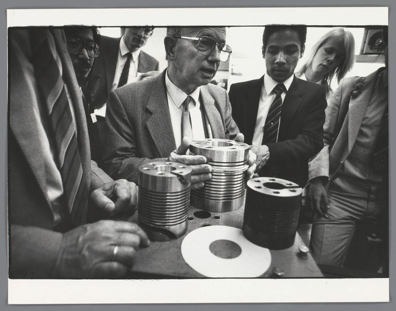 The image is a black and white photograph featuring several individuals gathered around the examination of various mechanical parts. The focus appears to be on an older man in glasses, who seems to be explaining or demonstrating something about these metal components that he holds with both hands. He wears a suit and tie, suggesting formality, possibly related to business or academia.

Surrounding him are at least five other individuals: two men appear engaged in the discussion — one directly facing us while another looks on from behind; there's also an older man beside them who is partially obscured by his glasses and hands. On the right side of the image stands a younger woman with blonde hair, her expression attentive as she observes or listens.

The mechanical parts held are cylindrical metal objects, possibly industrial or engineering components like bearings or bushings given their visible internal hole structures designed for shafts. Their polished surfaces reflect light differently than does the surface on which they rest—a large circular piece of what seems to be machinery with holes in its center.

All individuals wear professional attire such as suits and ties except for one woman, indicating a possibly corporate setting rather than casual or academic environment typically associated with student excursions abroad.

The background is indistinct but suggests an  [...]