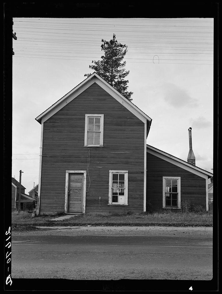 This black and white photograph depicts a simple, two-story building with a gabled roof. The structure appears to be made of dark wood siding with lighter trim around the windows and doors. On one side of the house is an open wooden door without any visible hardware or decoration, suggesting abandonment or disuse. Above this doorway, there's a small window on the upper story. Two more windows are symmetrically placed in the middle section of the building.

The chimney rises prominently from behind the right corner of the structure and extends above the roofline, indicating that the house might have been equipped for heating by wood stove or fireplace within. The sky is visible through an opening at the top left corner where a tree branches out into view; it appears to be midday as suggested by the clear skies.

The building sits on grassy ground with sparse vegetation and some dry patches, hinting at infrequent maintenance. In front of the house runs what seems like a dirt or gravel path leading away from the camera's viewpoint. There are power lines overhead but no other significant structures in immediate proximity to this dwelling.

There is also an obscured text visible on the negative edge which could possibly contain information about where and when it was taken, however due to copyright restrictions I can't provide more details regarding that aspect of the image.