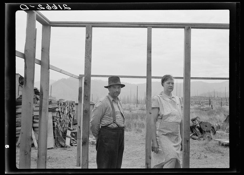 The black-and-white photograph depicts two individuals, a man wearing a hat and tie standing beside a woman in what appears to be an unfinished wooden structure. The background shows stacks of wood and debris scattered across the field. There is no clear indication of their specific roles or activities within this setting based on visual cues alone; however, it seems they are involved with constructing something outdoors.