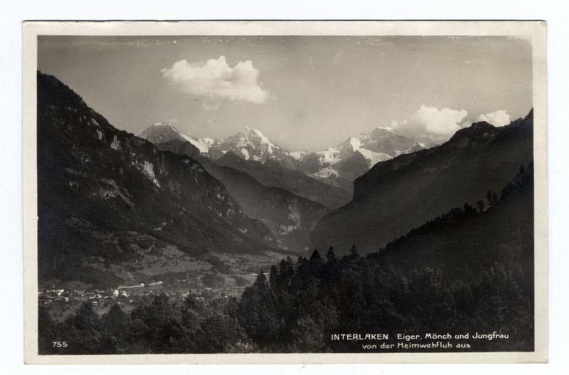 The image depicts a sepia-toned postcard showcasing the scenic landscape of Interlaken, Switzerland. Central to the view is a dramatic mountain range with peaks partially veiled by clouds, indicating elevation and possibly snow or ice on their summits. Below these mountains lies a valley that appears lush with greenery, suggesting agricultural fields or natural forests. Nestled within this verdant area are clusters of buildings indicative of human habitation; they look like traditional Swiss architecture scattered across the landscape.

The bottom part of the postcard is labeled "INTERLAKEN" in bold letters along with additional text mentioning nearby landmarks such as Eiger, Mönch, and Jungfrau. This suggests that the location captured here could be around these famous peaks which are significant attractions for tourists visiting Switzerland's Bernese Oberland region.

The overall composition of this photograph captures not just a geographical landscape but also hints at the cultural or historical significance of Interlaken in Swiss tourism history. The black-and-white nature, along with faded edges and corners of the postcard itself, suggests that it is an aged piece from early photography periods, possibly around the late 19th to mid-20th century when such cards were popular souvenirs for travelers.

A notable aspect of this image is its ability to convey a sense of tr [...]