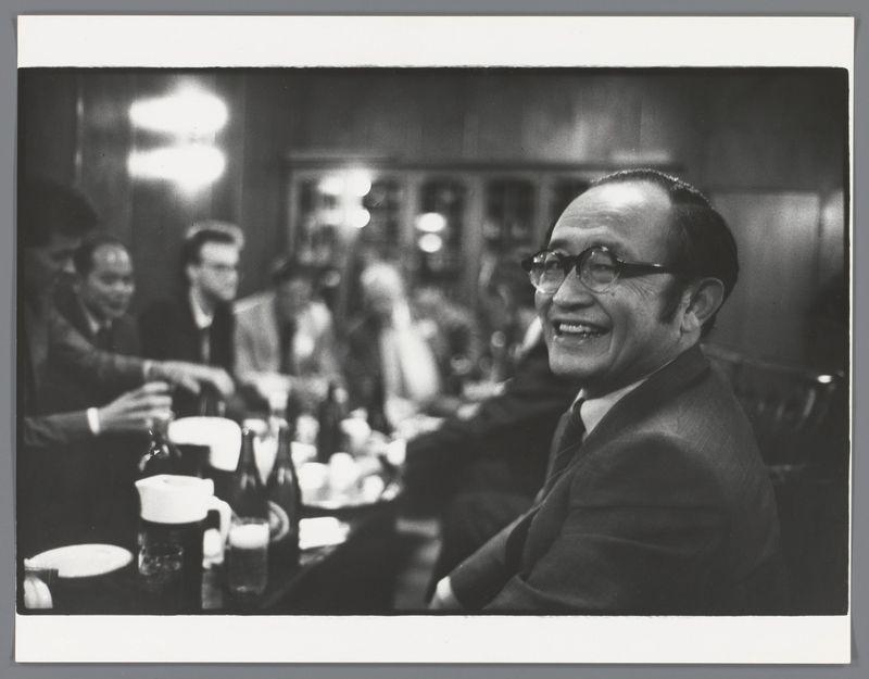 The image is a black and white photograph depicting what appears to be a social gathering in an indoor setting, possibly during the evening given the warm lighting. In the foreground sits a man who seems to be of East Asian descent with glasses, smiling broadly at something or someone outside the frame. He has dark hair receding from his temples, is wearing a suit and tie, suggesting formality for this event.

Behind him, there are blurred figures that indicate other people seated around tables covered in white tablecloths, which implies it's likely a dining establishment such as a restaurant or banquet hall. The attendees seem engaged in conversation; their hands suggest they might be gesturing during the discussion. There is no clear indication of any specific occasion from these details.

The photograph captures a candid moment, with its depth-of-field focused on the man closest to the camera and out-of-focus for those behind him. It's an intimate portrayal that hints at camaraderie or celebration among acquaintances or colleagues gathered together in this setting. The grayscale tones add a timeless quality to the scene.

There are no explicit indications of personal details such as names, occupations, or exact locations which would be pertinent given ethical considerations around privacy and data protection norms when describing images publicly accessible on platforms [...]