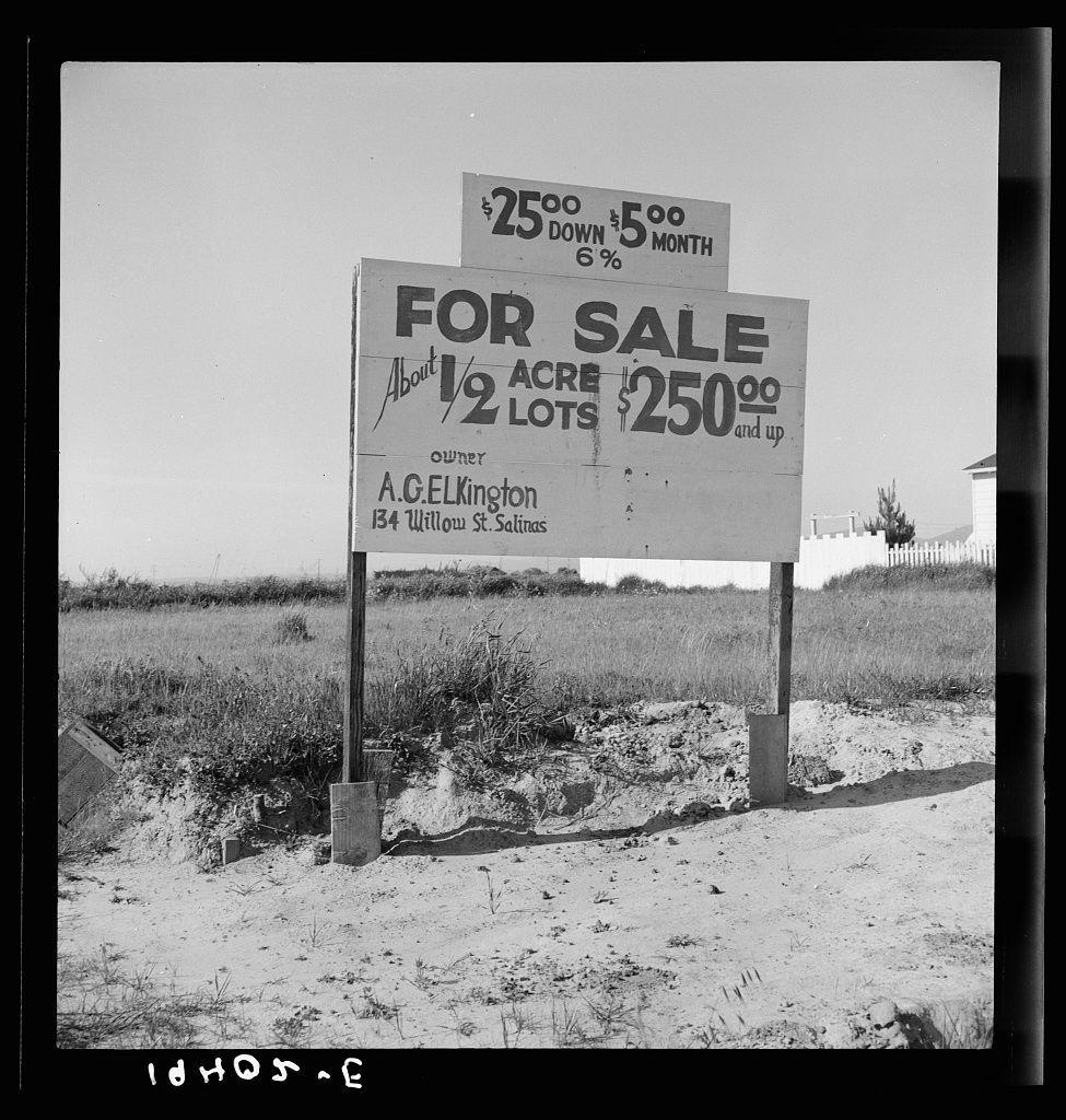 The image is a black and white photograph depicting two signs on an uneven sandy surface with sparse vegetation. The top sign, larger of the two, reads "FOR SALE" in bold capital letters at the center, indicating that property or land is being offered for sale. Below this main heading are details about what's available: approximately 1/2 acre lot size and a price listed as "$250 down $5oo/month." Further information specifies that it includes lots of "1/2 acre" with prices ranging from "$250" to an unspecified higher amount.

The second, smaller sign is positioned above the first one. It appears partially obscured but seems to provide additional details about a different aspect related to property or land for sale by A.G. Elkington at 134 Willow St., Salinas. The text on this part of the image also mentions "5% down" and "$500/month," suggesting another form of payment plan.

The background shows an open, flat landscape with what looks like a fence in the distance and some vegetation, possibly indicating that these signs are located near or within a field or undeveloped land. The lower part of the image contains handwritten text "1dHDS."E" which could be initials or code related to this photograph.

The overall impression is one of rural real estate advertisement with specific pricing information for potential buyers or investors interested in property development.