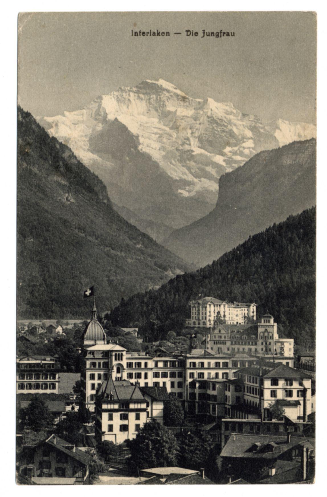 The image is a vintage black and white postcard featuring an early 20th-century European town nestled in the mountains. The foreground shows densely packed buildings with architectural styles indicative of late Victorian or Edwardian eras, including chalet-style homes, mansions, and what appears to be a hotel or public building prominently displaying a Swiss flag on its roof. Beyond this urban settlement is a vast expanse of rugged mountain terrain leading up to the Jungfrau glacier as indicated by text at the top left corner "Interlaken — Die Jungfrau." The mountains are majestic with snow-covered peaks, suggesting high altitude and possibly year-round cold temperatures in that region. There's dense forestation on either side of the valley where the town is situated, enhancing its secluded charm.