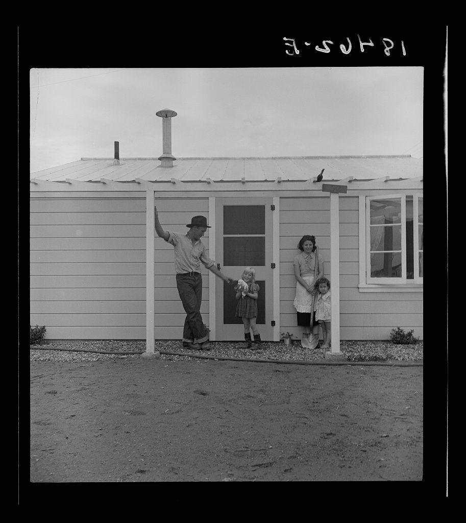 The image depicts a black and white photograph of three individuals standing in front of a one-story residential building. The structure appears to be made from light-colored horizontal siding, with two visible windows on the side facing away from the camera.

On the left, there is an older man wearing a wide-brimmed hat, suspenders, rolled-up pants, and what seems like work boots or shoes suitable for outdoor labor. He has his right hand raised above his head level in a relaxed manner while holding onto one of the door handles with his other arm extended towards it.

To the right side of the building stands an older woman wearing a dark-colored dress that extends down to her knees, along with darker shoes and socks. She is smiling gently at the camera, standing next to two young children who appear to be siblings due to their similar height and attire.

The first child on the left wears a short-sleeved floral-patterned sunsuit or sundress suitable for warm weather, complemented by knee-high boots. The second child stands slightly behind her sibling, wearing what appears to be a white dress with puffy sleeves. Both children are standing close together but not touching hands.

The ground in front of the building is unpaved and scattered with small stones, suggesting an outdoor setting that could potentially have been part of rural or semi-rural living conditions during the [...]