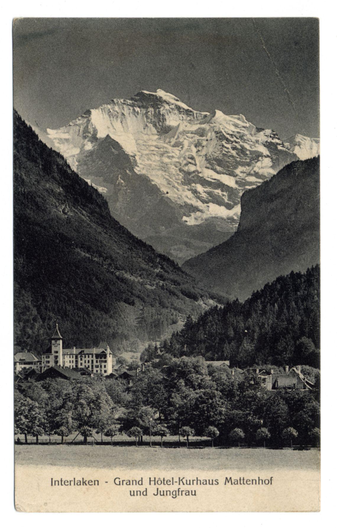 The image displays a vintage postcard featuring a dramatic mountain landscape with the prominent Jungfrau peak in the background. The mountains are covered with snow, indicating that it is likely taken during late autumn or winter. In the foreground and middle ground of the scene, there's an expansive view of lush greenery and dense forests on both sides flanking a valley where several buildings can be seen nestled into the landscape.

The most notable structure in this scenic tableau appears to be a grand hotel named "Grand Hôtel-Kurhaus Mattenhof," prominently featured centrally. Its architecture suggests it is quite large, with multiple levels featuring balconies and windows characteristic of European alpine resorts from past decades. Smaller structures are dispersed around the main building, likely serving various functions such as guest houses or service buildings.

The postcard carries German text at its bottom which translates to "Interlaken - Grand Hotel-Kurhaus Mattenhof und Jungfrau," indicating that this image is associated with Interlaken and the famous mountain known in English as the Eiger. This suggests a Swiss location, likely from a period when such hotels were popular for their role in alpine tourism.

The overall composition of the photo captures both the grandeur of nature's majesty and human attempts to create comfort within it through architecture, h [...]