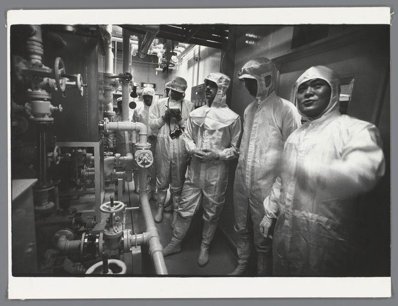 The image is a black and white photograph depicting several individuals in protective suits, possibly made of latex or another material. They are standing inside what appears to be an industrial setting with various pipes, valves, and mechanical equipment surrounding them. The people wear helmets that cover their heads completely except for the eyes which have visors, suggesting they might be working within a controlled environment where such protection is necessary. Some hold small objects in hand while others gesture or look at the camera.

The attire of these individuals suggests an emphasis on safety and cleanliness often associated with environments handling hazardous materials or requiring sterile conditions, like laboratories or chemical plants. The presence of multiple figures dressed alike indicates that they might be part of a team engaged in specialized work within this industrial setting.