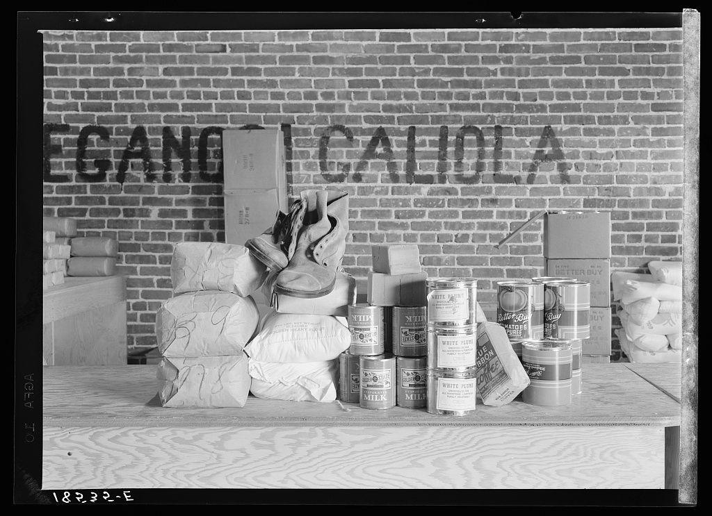 The image depicts a collection of food items and supplies arranged on what appears to be a table or counter against a brick wall with the words "GANG CALIOLA" painted onto it. The assortment includes several bags, cans, containers, and boxes that are likely part of emergency rations for agricultural workers in Bakersfield, California during times of destitution. This photograph is associated with the Farm Security Administration's (FSA) efforts to provide food and clothing grants to those in need amidst economic hardship.