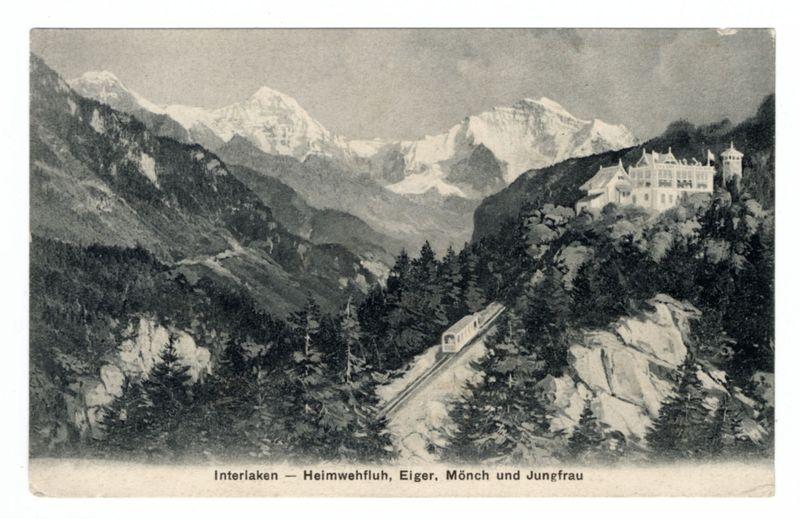 The image is a sepia-toned, vintage postcard depicting an aerial view of a mountainous landscape with railway tracks. There's a grand castle-like structure perched on the edge of a cliff overlooking the valley below. The mountains in the background have snow-capped peaks and rugged terrain. Forests are visible at various elevations across the scene, including dense clusters near the base of the cliffs where the train travels through.
The text "Interlaken — Heimwehfluh, Eiger, Mönch und Jungfrau" is printed below, likely indicating locations associated with this scenic view. The photograph captures a sense of early 20th-century transportation and tourism in Switzerland or Germany's Alps.