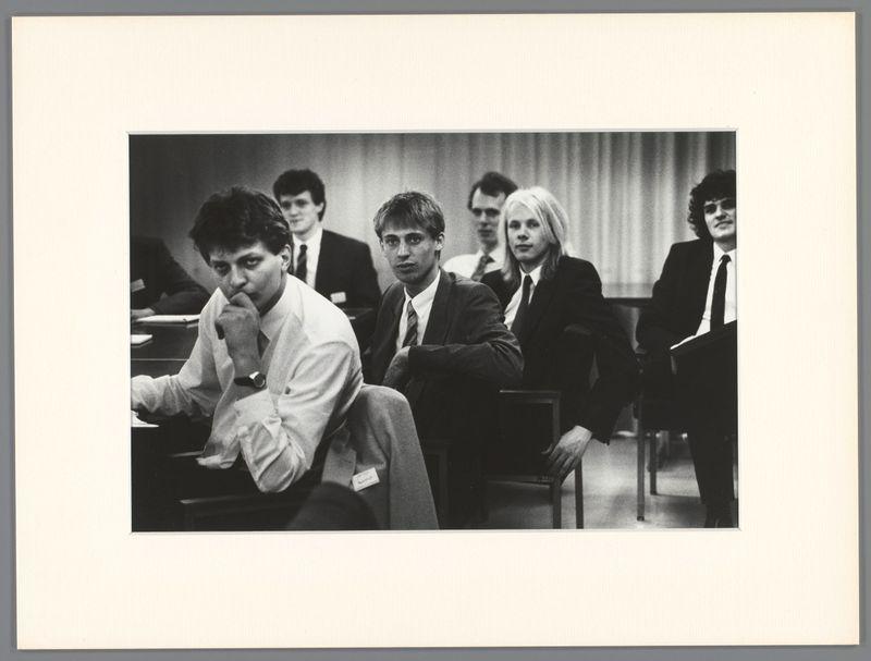 A black and white photograph showing a group of individuals seated in rows, appearing to be at an indoor gathering or meeting. The photo is framed with the image centered on a plain backdrop that could suggest it was taken within a room designed for events such as lectures or workshops.

The people are dressed formally; men wear suits and ties while one individual has their hair styled long, which indicates this may have been in the late 20th century. They show varied expressions: contemplative, engaged, attentive, with some looking directly at the camera and others glancing away thoughtfully.

There's a sense of engagement as if they are listening or reacting to someone speaking out of frame. The lighting is even, casting no strong shadows on their faces, which suggests an interior setting without direct sunlight or intense artificial light sources in use during this photograph. There appears to be text above the group but it’s not legible.

The image captures a candid moment within what seems like a professional environment and has artistic value due to its composition, lighting, and subject matter, indicative of work by Ed van der Elsken as per provided link information which suggests a cultural or historical context.