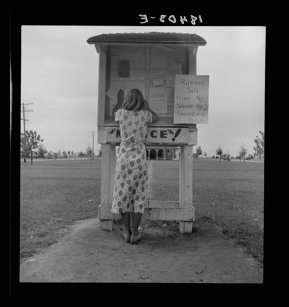 The image depicts a woman in mid-20th-century attire, standing and reaching into or touching the mailbox of what appears to be a communal bulletin board. She's wearing a patterned knee-length dress with closed-toe shoes. The structure housing this mailbox is labeled "SPECKLE," suggesting it serves as an information point within its community setting.

The background shows a vast open field, indicative of rural or suburban America during the time period suggested by her attire and hairstyle. There's also text visible on signs attached to the bulletin board: mentions of rummage sales with specific dates listed (Friday November 1st) along with some other information that is less clear due to resolution constraints.

The black-and-white tone of this photograph, alongside its monochromatic hues, points towards it being part of a photojournalistic series documenting aspects of daily life and community activities during the era. The image captures an everyday moment likely from rural America post-World War II when such communal bulletin boards were common for disseminating local information.

In terms of context provided by other references like "Untitled photo," this could be connected to communities where migrant labor was prevalent, potentially linked with agricultural regions in California or similar states that saw significant seasonal worker movement. This kind of communit [...]