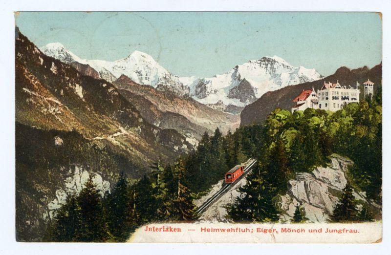 This image is a vintage postcard depicting the scenic beauty of Interlaken, Switzerland. The card shows snow-capped mountains in the background under a clear sky with shades ranging from light blue to hints of pink near the horizon. In the foreground, there's dense forestation indicative of mountainous terrain, featuring various conifer species and some scattered rocks or boulders.

A striking feature is an elevated railway track snaking its way through the landscape, leading towards a large white building with red roofing at the summit. The train on this narrow-gauge railroad appears to be in motion, enhancing the sense of travel and adventure associated with visiting such remote areas during that era. 

Text inscribed across the bottom reads "Interlaken—Heimwehfluh; Eiger, Mönch und Jungfrau," which translates from German as "Interlaken—the homesickness for; Eiger, Mönch, and Jungfrau." These are renowned peaks in Switzerland's Bernese Alps. The mention of these mountains on the 