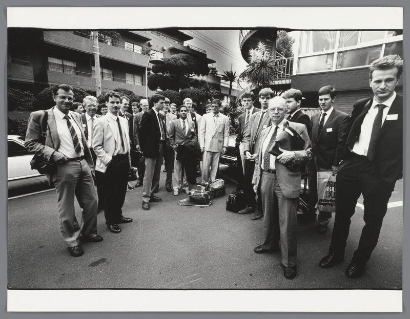 This black and white photograph depicts a group of individuals, presumably students or academics given their attire—suits, ties, and formal clothing. They are gathered outside what appears to be an urban residential area with multi-story buildings in the background featuring balconies and windows. The presence of luggage on the ground suggests they might be traveling or have just arrived at this location.

The group is diverse in terms of age and possibly nationality; some individuals display features that suggest European descent, while others appear different—indicating a multicultural mix. Their expressions vary from smiles to more neutral or serious looks, with most looking directly into the camera's lens. The image captures what seems like an organized event or outing.

Additional information about this photo can be found through the provided link: Groepsportret van Groningse studenten en docenten in Japan (Group portrait of Groningen students and lecturers in Japan) by Ed van der Elsken, dated between 1985-1990. This suggests that these individuals may have been part of a delegation or exchange program with Japan during this period.

This photograph is significant as it provides insight into the international relations and exchanges occurring among students from different backgrounds in Europe at least around those years. It highlights cultural diplomacy, educationa [...]