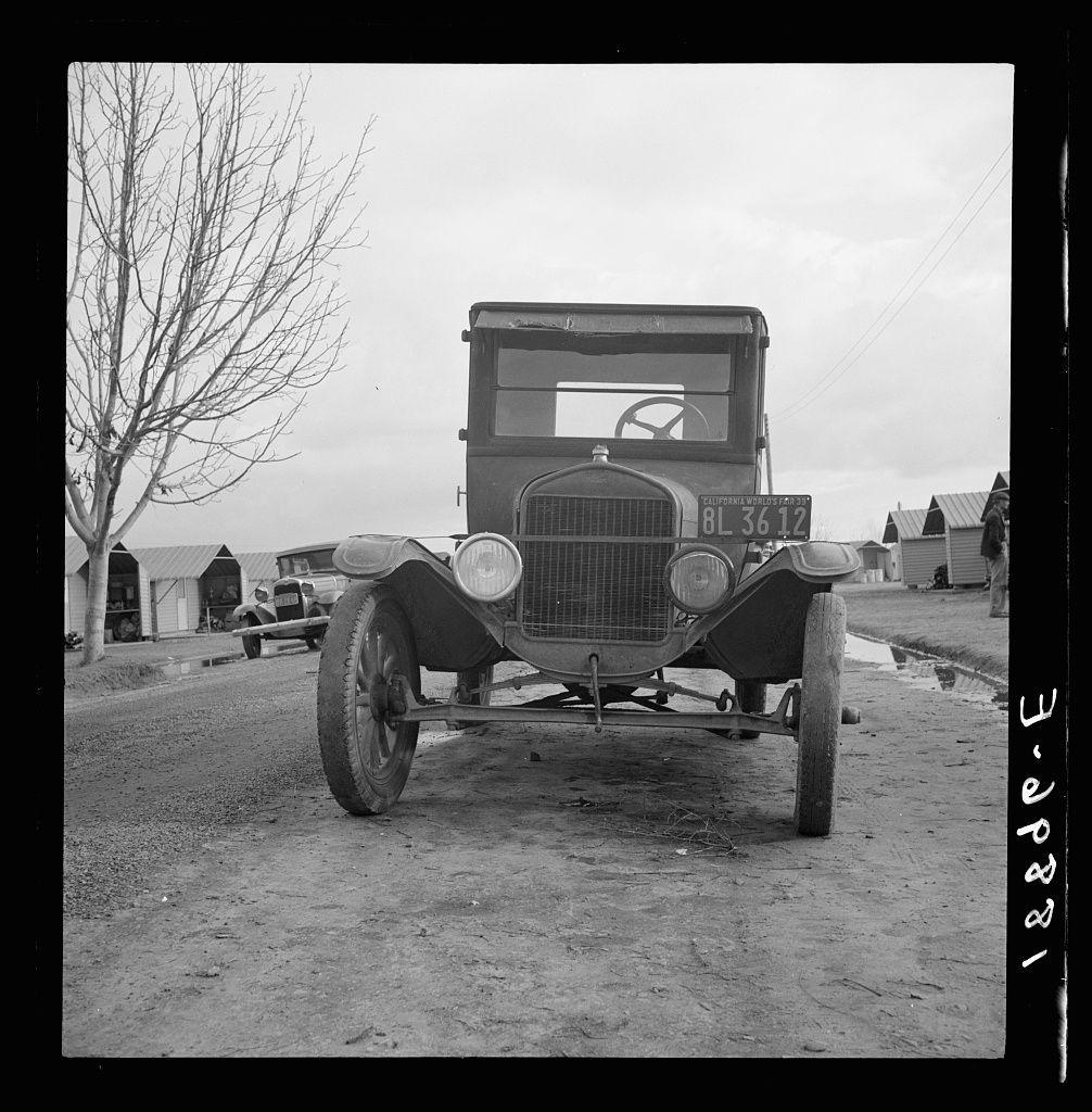 The image displays a black and white photograph of an antique Model T Ford truck on what appears to be unpaved, possibly rural roads. The vehicle is facing the camera directly, centered in the frame with its front clearly visible showcasing circular headlights, a distinctive grill design characteristic of early 20th-century automobiles, and large, round wheels indicative of that era's technology.

In the background, there are several structures resembling farm sheds or outbuildings, suggesting an agricultural setting. A barren tree stands to the left side in the foreground on what appears to be muddy ground with puddles indicating recent rain. To the right, two individuals can be seen walking away from the camera; one is wearing a hat and seems to have rolled-up sleeves.

In terms of context or background story based on this image alone without additional information provided by external resources such as "Untitled photo," it might represent an early 20th-century setting in America. The Model T Ford was famously known for its utility, affordability, and accessibility during that time period. Its presence could be indicative of the mobility and economic conditions of rural or semi-rural areas around this era.

The image is part of a collection potentially linked to Farm Security Administration (FSA) migratory labor camps in Farmersville, California as mentioned by another  [...]