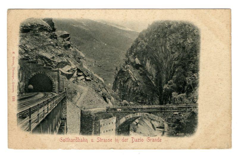 The image depicts a vintage black and white postcard featuring an old railway through a mountainous landscape. The railway is shown in two sections: on the left, a train emerges from a tunnel carved into a rocky cliffside, with tracks extending further back out of sight; to the right, another section of track bridges over a valley via stone arches supported by cylindrical pillars. This bridge passes above what appears to be an ancient building partially hidden behind foliage and rock formations. The terrain on either side features rugged mountainside slopes covered in sparse vegetation, indicating steep and possibly challenging geography for construction.

The image bears inscriptions that provide historical context: "Gothardbahn u. Strasse in der Dazio Grande" is printed at the bottom of the postcard, which translates to "Gotthard Railway and Road through Dazio Grand." A date stamp reading "1927/36" suggests when this photograph or card was produced or used.

The overall impression evokes a sense of early 20th-century engineering achievements in overcoming formidable natural barriers. The postcard's aged appearance, with its slightly worn edges and discoloration at the corners, enhances the historical ambiance of the scene captured here.
