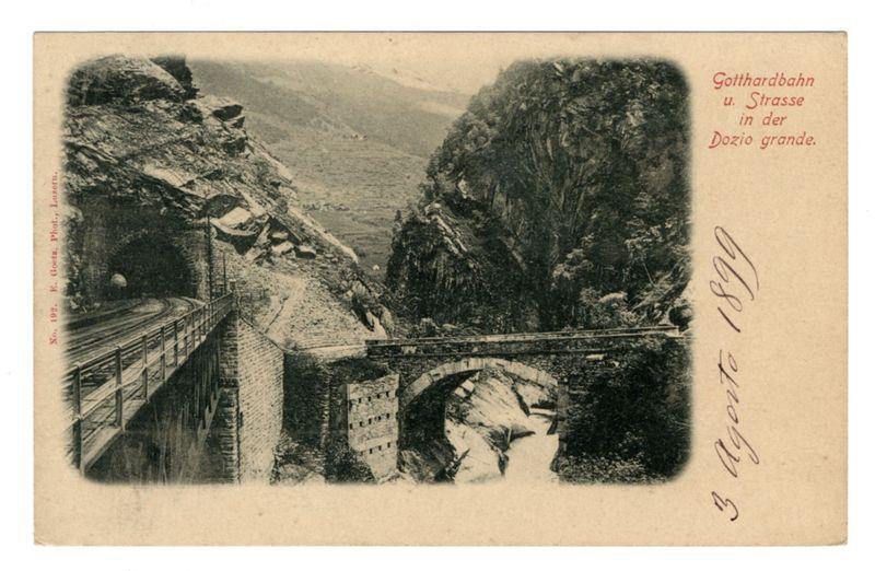 The image depicts an old, monochromatic postcard featuring a vintage scene of a railway bridge crossing over a mountainous landscape. On the left side, there's a stone tunnel cut into the hillside leading to another structure that appears to be part of the train tracks or infrastructure related to it. Adjacent to this is a wooden pedestrian bridge with railings, connecting both sides of what seems like a ravine.

The central focus of the image is an arched railway bridge built over a riverbed surrounded by rocky terrain and vegetation on either side. The background showcases mountainous regions, indicating that the location could be in a valley or gorge.

At the top right corner of the postcard, there's text written in German: "Gotthardbahn u. Strasse in der Dözio grande," which translates to "Gotthard Railway and Road through the Grand Dōzio." This suggests that the scene is captured near a pass or gorge named after its Italian equivalent 'Grand Dōzio'. Below this, there's handwriting on the postcard with a date written as "3 Agosto 1826," which translates to August 3, 1826. The number and name of the photographer are also inscribed at the bottom left corner: "No. 1904. E. Goltz Phot., Lucerne."

The overall condition and style indicate that this postcard is from an earlier period in photography history, possibly late 19th to early 20th century. The signature on the card [...]