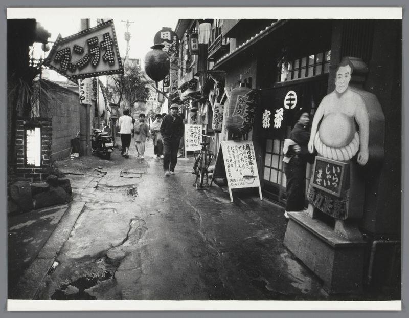 The image is a black and white photograph depicting an urban street scene. The focus of the composition appears to be on a bustling alleyway with various signs, storefronts, and people walking along the sidewalk.

On the left side, there's a large signboard hanging from a structure above that features Japanese characters, suggesting it might indicate the name or nature of a business establishment in this area. Below the signboard is another smaller rectangular sign attached to what seems like an old-style lamp post with similar text and symbols indicating some commercial activity.

In the center foreground stands a life-sized statue of a sumo wrestler on top of a pedestal, likely serving as an advertisement for a nearby restaurant or business related to Japanese cuisine. The statue has large round features characteristic of traditional representations in Japan's sumo culture.

Several individuals are visible walking down the street; one person appears prominently engaged with some sort of equipment possibly connected with food preparation. There is also another figure closer to the camera, holding what seems like a folded umbrella or similar object over their head while looking towards something out-of-frame on the right side.

The ground shows signs of wear and age, with patches where the concrete has chipped away revealing underlying materials beneath it. The street its [...]