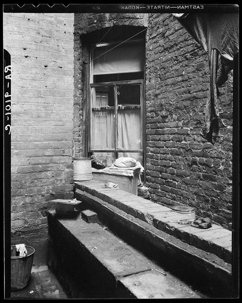 The image depicts a dilapidated outdoor area of an old brick building. The focus is on the side of what appears to be Mr. and Mrs. Jacob Solomon's tenement dwelling located at 133 Avenue D, New York City as mentioned in context provided from https://images.loener.nl/DorotheaLange/full/6495/6495b844ba25398d90c5038a.jpg. The view shows a narrow alleyway with worn brick walls on both sides, leading to an open window above which curtains are partially drawn down.

On the left side of the frame is a bucket filled with discarded items and hanging laundry from a line strung across two poles outside the window. To the right stands another pole holding up more laundry in front of the window. Below these elements lies a stone step leading to an outdoor ledge, where various objects such as buckets, containers, and possibly personal belongings are scattered.

The overall impression is one of neglect and poverty, reflecting the living conditions during that period. The black-and-white tone adds to the historical context and somber mood of the scene.