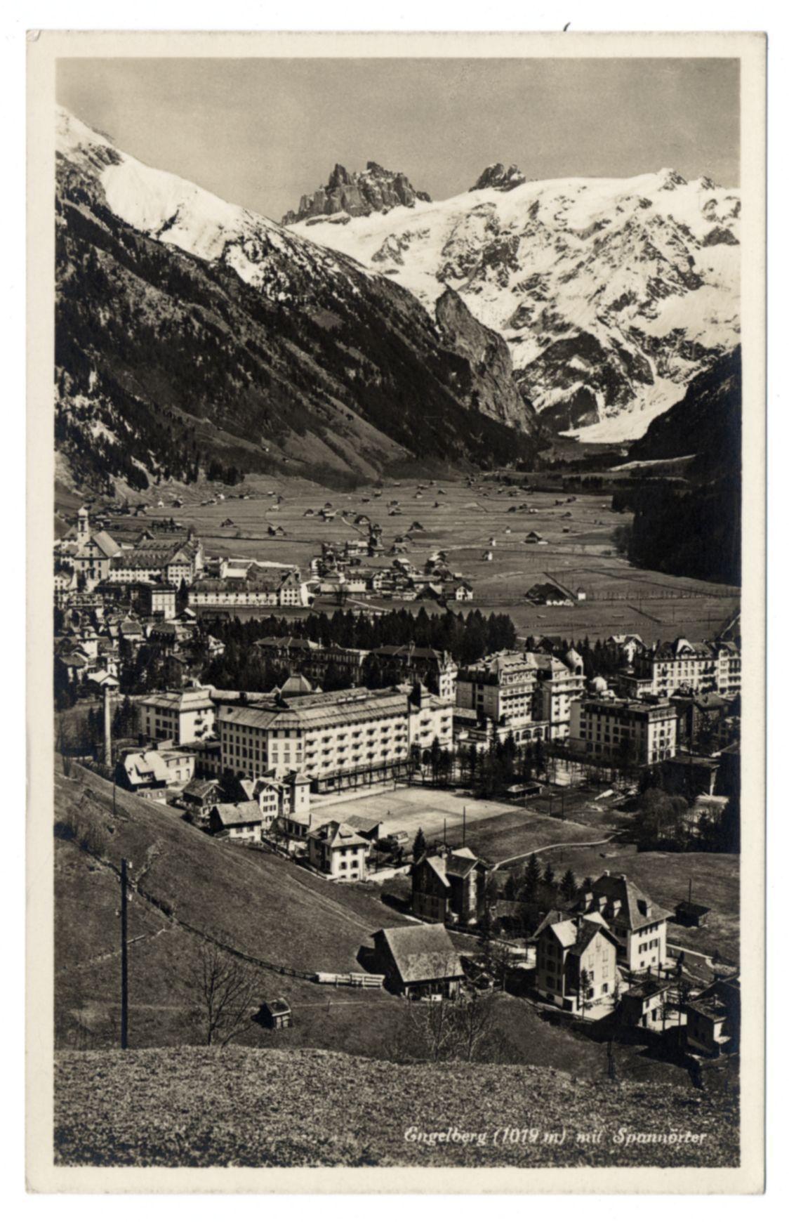 This black and white photograph depicts a quaint Swiss village nestled in the mountains. The scene is dominated by expansive, snow-capped peaks that loom dramatically over the valley below. Nestled amidst these towering natural sentinels are multistory buildings with traditional architecture indicative of European alpine villages. A railway line snakes its way through the landscape, adding another layer to this picturesque tableau.

In the foreground lies a vast expanse of fields and cultivated plots stretching towards the village's central area where clusters of residential structures dominate. The contrast between the rural farming grounds and urban development is stark yet harmonious.

Among these buildings stands an imposing hotel or similar establishment with multiple stories, likely serving as accommodation for tourists exploring this Alpine locale. This building’s size suggests its significance within the community.

The image appears to be a historical photograph given its monochrome tone, which lends it a timeless quality. It captures not just the physical environment but also hints at the lifestyle and culture of villagers during the era in which it was taken. The absence of modern infrastructure indicates that this village maintains traditional ways amidst an ever-changing world.

In terms of text within the image, "Gingelberg (1019 m) mit Spanower" is inscribe [...]