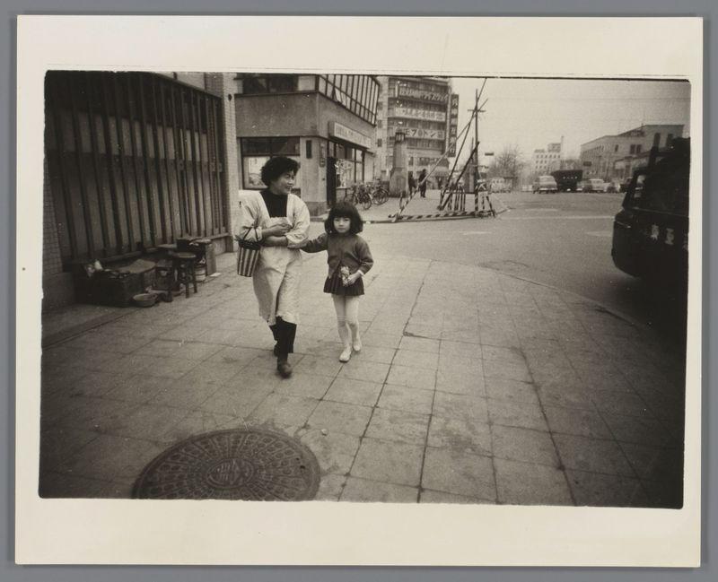 The image depicts a street scene, captured in black and white. It features an adult woman leading the way while holding hands with a young girl who appears to be walking beside her or slightly behind. They are on what looks like a paved city sidewalk. The surrounding environment includes urban elements such as buildings, windows, a manhole cover, a parked motorcycle with sidecars in the background, and some debris alongside the curb.

The adult woman is wearing boots, pants, a jacket over her shoulders, and appears to be dressed for cooler weather or perhaps an occasion that calls for more formal attire. The young girl has on what seems like school uniform trousers paired with a dress top and white leggings, carrying something in one hand while holding onto the adult's arm.

In terms of context provided by the caption "Vrouw loopt met meisje op straat," which translates to English as 'woman walking with girl on street,' it suggests that this could be a candid moment captured during everyday life. The woman and child seem engaged, possibly in conversation or simply sharing an intimate walk through urban Japan around 1959-60.

The photograph is presented within what appears to be the frame of a Polaroid instant camera print, which adds a vintage feel to this historical snapshot from Ed van der Elsken's collection.