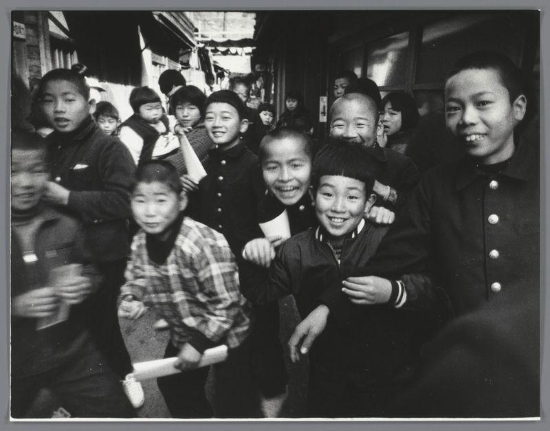 The image is a black and white photograph showing a group of people, presumably children based on their size and features. They are gathered closely together, with many smiling at the camera or making playful faces, suggesting a moment captured during a casual outing in an urban environment.

In the foreground, there's a child to the left holding what appears to be a rolled-up paper or map, another pointing towards something off-camera on their right side; behind them stands a figure wearing a dark coat with buttons. The central area of the image is populated by several children who seem excited and joyful, with one in particular smiling broadly at the camera.

Behind this group are other individuals whose faces are partially obscured but appear to be partaking in similar activities or waiting their turn for something out of frame. Their attire includes various styles indicative of a colder climate; many wear coats, hats, scarves, and gloves suggesting it's possibly winter.

The background shows elements typical of an urban street setting such as shops with visible signage, hanging items that could be clothes drying on lines, or other merchandise being displayed for sale. The scene conveys a sense of community and everyday life in Japan during the late 1950s to early 1960s.

The photograph carries historical significance; it's associated with Ed van der Elsken (died 2005) [...]
