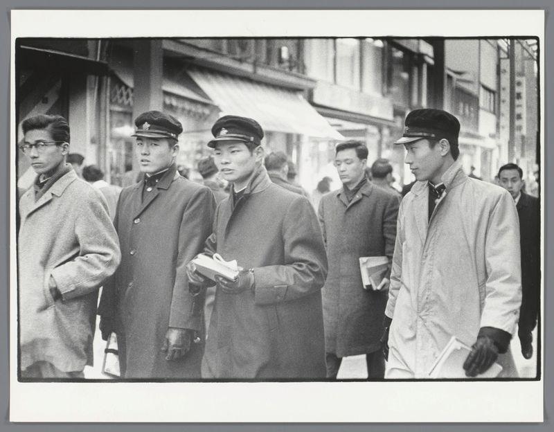 The image is a black and white photograph featuring five men walking on what appears to be a busy street. The central figures are two middle-aged Asian males in military-style caps, one of whom carries an item that could possibly be a box or package. To their left stands another younger male with glasses wearing a light-colored jacket over a dark coat. On the right side is yet another young man dressed similarly to his companion on the opposite end – both are clad in coats and seem engrossed in walking.

The background suggests an urban environment, potentially Japan based on historical context provided by external resources (as indicated at the bottom). The men exhibit expressions of attentiveness or possibly concern. Behind them is a blur of other individuals who could be pedestrians going about their day, indicative of bustling city life during that era.
