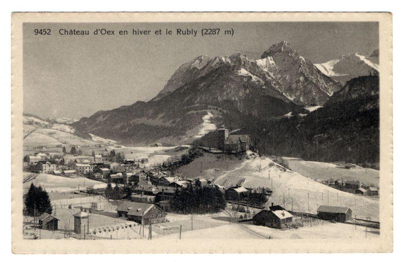 The image shows a black and white vintage postcard depicting a snowy landscape with mountainous terrain in the background. The caption on the top of the card reads "9452 Château d'Oex en hiver et le Rubly (2287 m)" which translates to "Château d'Oex in winter and Le Rubly (2287 meters)." Below this text is a view of a small village nestled among snow-covered hills. The town has various buildings, some with sloped roofs covered in snow, indicative of the cold climate typical for such regions during wintertime.

In the foreground, there's an expanse of open ground that appears to be partly cleared from the snow, possibly used as fields or pathways. There are a few bare trees scattered around and wooden fences bordering some properties. A ski slope can be seen on the left side leading up towards the mountains, with several individuals visible at various points along its incline.

The postcard carries signs of age; it's slightly worn out with creases indicating that it has been handled or stored in less than optimal conditions over time. The overall impression is one of a quaint and picturesque winter scene from a bygone era, capturing the charm of life amidst alpine mountains during snowy months.