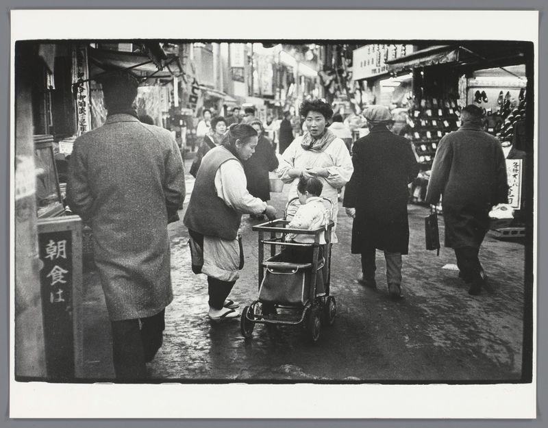 The image captures a street scene in Japan, likely from the late 1950s to early 1960s based on clothing and environment. A group of people is seen walking down what appears to be an outdoor market or shopping area with various stalls along the sides. In the center foreground, there's a person dressed in traditional Japanese attire attending to someone seated in a small cart-like chair, suggesting they may need assistance due to mobility issues such as age or disability.

The surrounding individuals seem engaged in their activities; some walking away from the camera and others interacting with each other or focusing on market goods. The photograph is monochromatic, typical of that era's street photography, which adds a sense of historical context. Notable features include the wet ground reflecting light, indicating recent rain, and traditional signage visible along the streets.

The black-and-white tone emphasizes textures such as clothing fabrics, pavement dampness, and metallic parts on carts or bicycles. The overall atmosphere is one of everyday life with an undertone of community support amid urban bustle. Additional information about this image can be found in Straatbeeld met baby in kinderwagen, Japan, Ed van der Elsken, 1959 - 1960 on the specified link provided for more details and context regarding the photographer's work during that period.