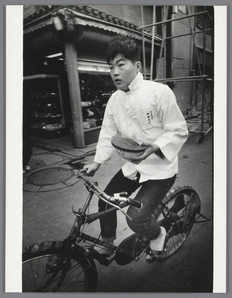The image depicts a young man riding a bicycle on what appears to be an urban street at night. He is wearing a white chef's jacket with Chinese characters embroidered in black, suggesting he may work as a cook or food delivery person. In his right hand, which is extended outward from the handlebars of the bike, he holds a plate and bowl that are used for serving food. The man has short hair, dark eyes, and appears focused on something ahead of him, possibly the street or destination where he's delivering the meal.

The bicycle looks sturdy with thick tires, commonly associated with city bikes designed to navigate through various urban terrains. In his left hand, which is gripping one of the bike handles, there seems to be a bag hanging from it, likely used for carrying additional items while on duty as a food delivery person. The background features elements typical of an industrial or maintenance area such as scaffolding and what appears to be construction materials.

The photograph has a high-contrast black-and-white look which adds a sense of timelessness and captures the essence of nighttime city life, emphasizing the subject's journey through his workday in Japan during 1959 - 1960. The image is part of an archive by Ed van der Elsken, as indicated by the provided link with additional information about this specific photograph.