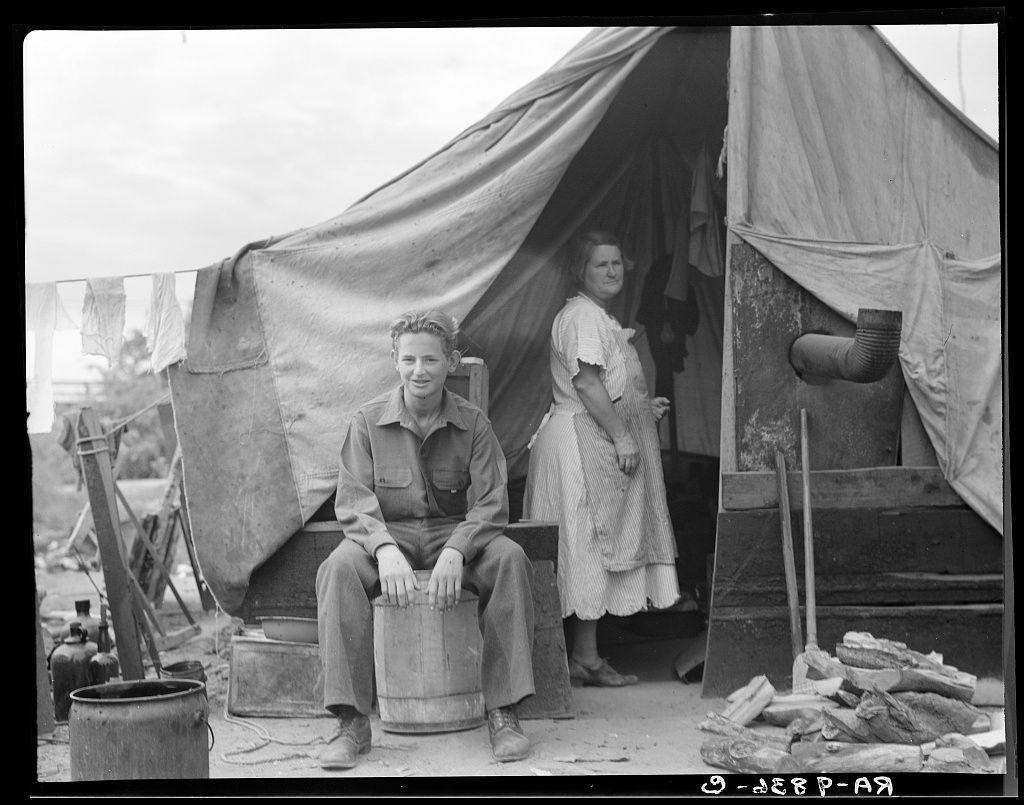 A black and white photograph depicting a family encamped near Porterville, California. One individual is sitting on a wooden barrel in front of a makeshift tent with various items such as firewood, clothes hanging to dry, and storage containers scattered around the area indicating an outdoor living situation during migration or hardship period likely connected to agricultural work like orange groves based on historical context.
