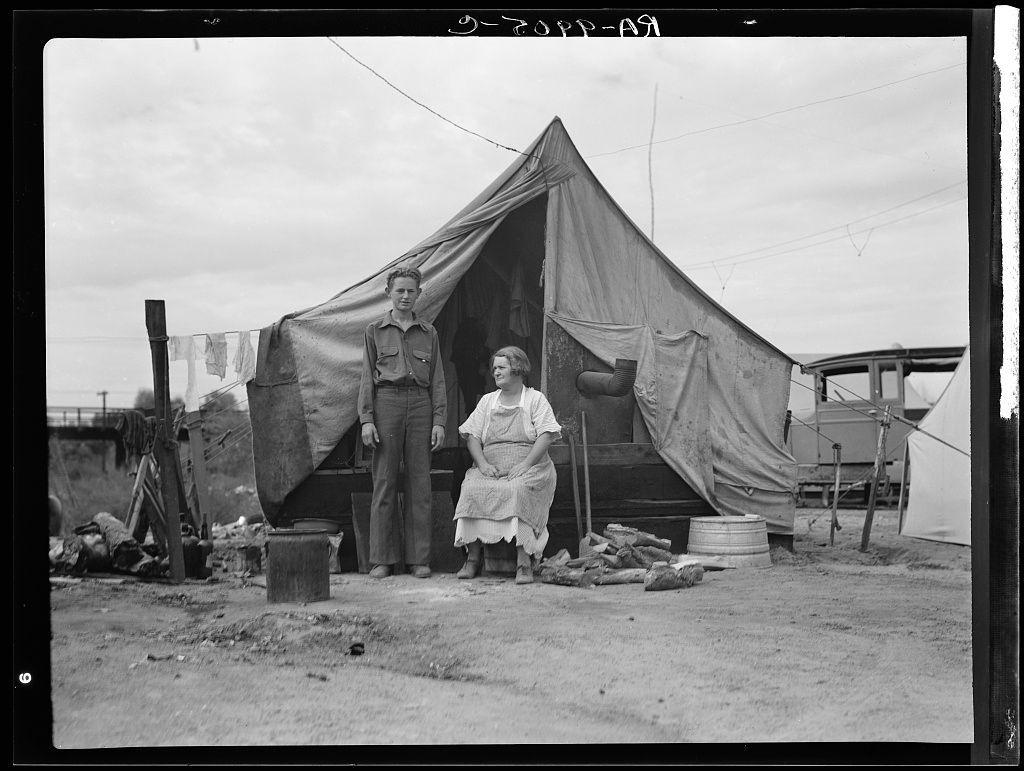 The black and white photograph shows a migrant family of five, with two individuals seated in front of a makeshift tent. The boy is on the left side wearing rolled-up pants and an open-sleeved shirt, while the woman appears to be older, possibly his mother or aunt. She wears a dress typical for that era, indicating modesty and practicality suitable for manual labor.

The living conditions are stark—there's no bed; instead, they sit on what seems like rags. The ground is unpaved dirt with some scattered items around: buckets, wood logs suggesting makeshift firewood collection or chopping activities, a barrel likely used as storage or possibly fuel container. A metal object resembling an oil drum could be for storing cooking oils or other liquids.

The tent material appears to have been repurposed and patched up—a reflection of their transient lifestyle. In the background lies more encampment structures indicative of larger community living arrangements typical during Depression-era rural migrations, particularly in agricultural areas like California's Central Valley known for its orange groves which were heavily dependent on migrant labor.

The overcast sky contributes to a somber tone and may reflect weather conditions or economic gloom. Despite the hardships implied by their surroundings, they exude an air of resilience and adaptability—key traits essential during that e [...]
