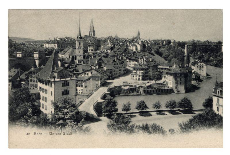 The image depicts a vintage black and white postcard showing an aerial view of Bern, specifically the "Untere Stadt" area. The photograph captures a cityscape with dense clusters of buildings featuring steeply pitched roofs characteristic of traditional Swiss architecture. Notable structures include churches or towers that rise above the surrounding rooftops, suggesting their prominence in the urban skyline.

In the foreground, trees and greenery are visible along what appears to be a riverside promenade, leading towards water bodies indicative of rivers flowing through Bern. The scene is framed by rolling hills in the background under a clear sky, adding depth to the panoramic view. A bridge spans across one river section on the right side.

The photograph bears handwritten text at its bottom edge and printed captions indicating "48" possibly representing an edition or series number and "Bern — Unterer Stadt," specifying the location within Bern's urban layout. The image conveys a