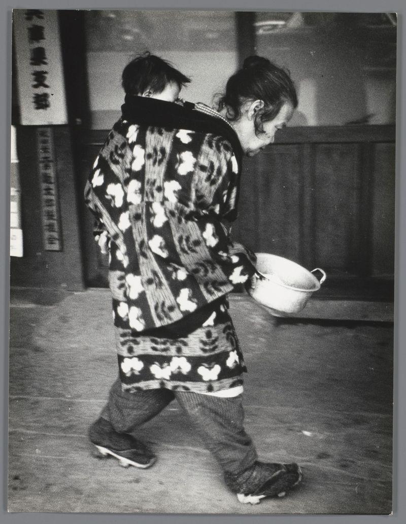 The image depicts a black and white photograph of two individuals walking on what appears to be an urban street. The person in the foreground is wearing traditional Japanese clothing, specifically a patterned kimono with large floral motifs, which contrasts against their dark pants and shoes. They are carrying a bowl or pot under one arm as they walk forward.

The second individual stands behind partially obscured by the first but also appears to be walking on the street. The background includes an entranceway with signage that has Japanese text above it, suggesting this scene is taking place in Japan during the late 1950s-early 1960s as indicated by the reference information provided.

The photograph captures a candid moment of everyday life and reflects cultural aspects from its time. The attire and setting give insights into daily practices or activities common to that era, potentially relating to street food vending or similar urban customs in Japan during those decades.