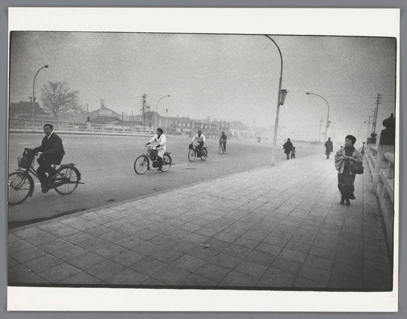 The image depicts a street scene, likely from the late 1950s or early 1960s based on its black and white presentation. Multiple individuals are seen traveling by bicycle along a road that appears to be in an urban setting with buildings visible in the background partially obscured by fog or mist.

On the left side of the street, two cyclists ride one after another; both wear dark clothing and helmets for safety. Further down the right-hand sidewalk, pedestrians walk: three individuals are seen walking away from the viewpoint, while a fourth walks towards it carrying what appears to be a briefcase over their shoulder.

The atmosphere is somewhat subdued with muted tones due possibly to weather conditions like fog or pollution which adds texture to this otherwise clear scene of urban life. The road itself seems relatively uncluttered and devoid of heavy traffic at the moment captured by this photograph, suggesting it might have been taken during a quiet period in Japan's streets.

