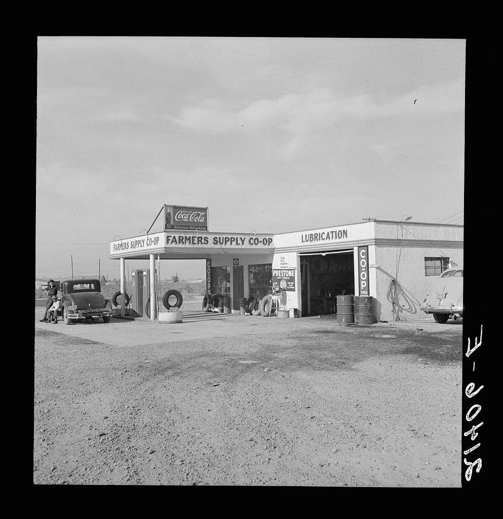 The image is a black and white photograph of an old farmers' supply co-op. The building features several signs, including one for Coca-Cola and another that reads "Farmers Supply Co-Op." There are also additional labels such as "Lubrication" and the word "Coop" prominently displayed on different parts of the structure.

In front of the building, there is a vintage pickup truck with two visible tires resting against it. A person can be seen standing beside the truck, seemingly engaged in an activity or conversation near one of the tire stacks. Another vehicle is parked to the right side of the image, and various equipment such as barrels labeled "Gas" are scattered around.

The ground appears dry and dusty, indicating arid conditions typical for agricultural areas like a farmers' co-op might be located. The sky in the background looks cloudy but not overly dark or stormy. Overall, this photograph captures a moment of rural life, likely from mid-20th-century America, reflecting on the everyday workings of such an establishment and its role within the community it served.