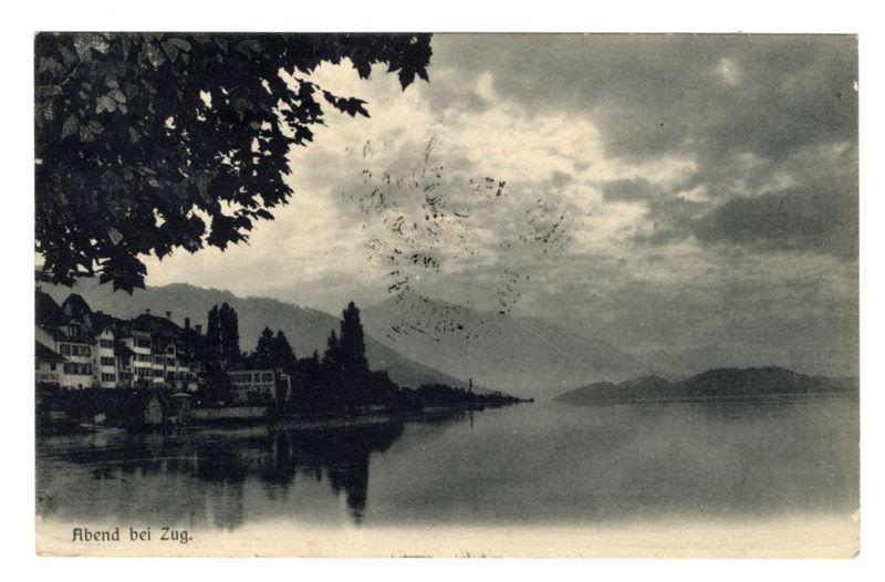 The image depicts a vintage black and white postcard showing a serene lakeside view. A body of water reflects the sky above, which is partly cloudy with some clearer areas where sunlight can be seen piercing through.

On the left side of the card, silhouetted trees appear in sharp contrast against a lighter backdrop, their dark branches extending towards the top edge and overhanging part of the frame on the right. Below this tree line lies a row of European-style buildings with multiple stories, characterized by white walls and brown roofs. Some have visible windows that hint at residential or commercial use.

In the center to right portion of the image, calm water stretches across the view towards distant mountains in silhouette against the sky. Several small islands break up the expanse of the lake. The landscape suggests a tranquil evening atmosphere with stillness emanating from both natural and human-made elements.

At the bottom left corner of the card is text that reads "Abend bei Zug," translating to "Evening at Zug" which indicates the location captured in this scene, likely referring to Zug, Switzerland.