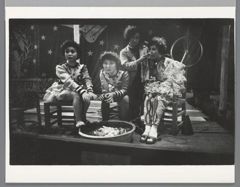The image captures a moment featuring four women dressed in costumes, seated on what appears to be a stage. The setting has a vintage feel with black and white tones that suggest the photograph may have been taken some decades ago.

From left to right: 
- A woman wearing a striped top is seated comfortably with legs crossed.
- Next to her sits another woman donning a traditional outfit, possibly indicative of Japanese culture given its style. She's engaged in holding a pair of chopsticks and appears focused on something out of frame.
- The third individual, centrally located within the group, seems slightly turned towards their companion on the left but is not directly interacting with them or anyone else at that moment.

The fourth woman to the right exhibits an expressive posture; she sits with one leg draped over another in a relaxed manner. She's holding what looks like a cigarette and appears animated as though conversing or making a gesture, adding dynamism to her characte