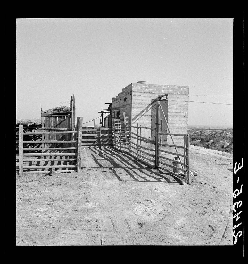The image depicts a rural scene with dilapidated wooden structures. In the foreground, there is an open gate leading to what appears to be fenced-off areas made of wood and brick. The ground shows signs of wear, possibly dirt or sand. A water jug stands next to one structure in the middle distance, indicating some usage despite the overall neglected state of these buildings.

The photograph's black-and-white color scheme suggests it is historical, as does its grainy texture and style. It has a handwritten label "ST-32-E" on the right side, likely used for cataloging or archival purposes during Dorothea Lange’s time working with her photographic documentation efforts in various regions.

This image captures an atmosphere of abandonment and decay, possibly indicating economic hardship or disuse at this location which may have been a part of rural livelihoods.