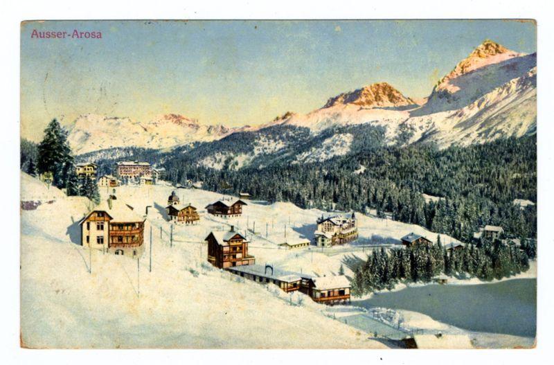 The image depicts a picturesque winter landscape, showcasing a quaint village nestled in the snowy mountains. The foreground features a collection of chalet-style buildings with steeply pitched roofs and large windows, indicative of alpine architecture designed for snowfall protection. These structures are scattered across gently sloping terrain blanketed in white snow.

In the midground, more substantial hotel or resort buildings stand out due to their size and prominent placement within the village layout. They appear to serve as a hub for visitors seeking refuge from the cold amid the idyllic setting. 

The background is dominated by towering mountains with snowy peaks that rise majestically against a clear sky. The slopes of these mountains are adorned with patches of evergreen trees, suggesting they may be populated during warmer months or higher altitudes where conditions prevent snow cover.

A lake can also be seen on the right side of the image, partially obscured by clus