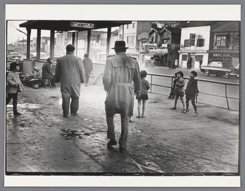 The image depicts a black and white street scene, likely from the mid-20th century. It shows several people walking on what appears to be a wet pavement. In the foreground, there's an individual in overalls with their back turned toward us; they seem to be leading or accompanying another person who is out of focus behind them.

In the middle ground, we see two small children holding hands and walking away from our perspective towards some buildings on the right side. To the left, a young girl wearing a coat walks alone down the street. In front of her stands an adult in winter clothing. There's also another person visible further back on the pavement.

On the upper part of the image, we see parts of what looks like market stalls or small shops with people moving about. The background features buildings and parked cars indicative of a busy urban environment during this period. A bus stop shelter is noticeable under which more individuals seem to be gathered out of focus.