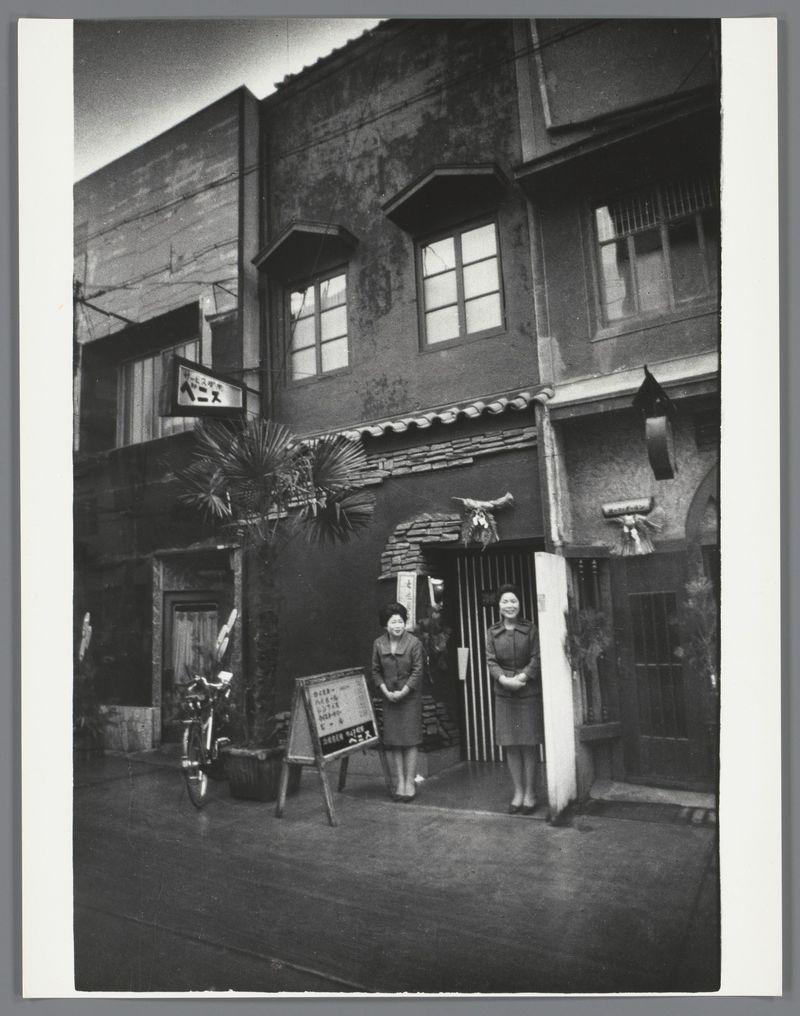 The image captures a monochrome street scene, likely from the mid-20th century based on architectural and fashion elements. It depicts two women standing in front of an establishment that appears to be a bar or restaurant. The building is aged with visible wear and features traditional Japanese design elements such as wooden beams and tiles along its eaves.

To the left stands a woman, possibly Asian, wearing a dark coat and pants, smiling at the camera. Next to her is another individual in similar attire but not clearly identifiable due to blurriness or angle. The entrance has a signboard with Kanji characters indicating it's an establishment open late (around 3am). A mannequin dressed in Japanese traditional garb stands guard outside.

A bicycle leans against the building, suggesting this was perhaps a common mode of transport at that time. There is also signage on various parts of the structure and what appears to be part of scaffolding or construction visible above it.