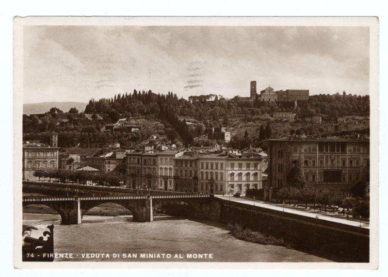 The image depicts a sepia-toned photograph of the cityscape in Florence, Italy. It shows an elevated perspective overlooking a river with a bridge crossing it and buildings sprawling down towards its banks on either side. The architecture is dense and historic, indicative of European urban design from past centuries.

A series of trees partially obscure some structures near the center background, indicating lush greenery or hillside terrain rising behind them. In the far distance sits a large castle-like structure atop what appears to be a hill with vegetation clinging to it. A clear sky spans above the landscape, though clouds are visible in patches across parts of the image.

In the foreground on the bottom left corner is text that reads "74 - FIRENZE- VEDUTA DI SAN MINIATO AL MONTE" which translates from Italian as 'Florence - View of San Miniato at Monte.' This suggests the title or location name associated with this photograph. The overall impression conveyed by the image capt