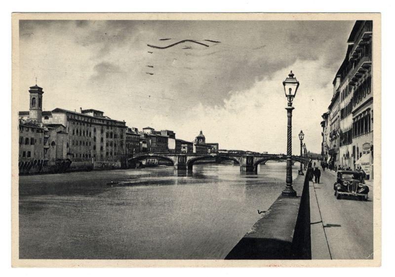 The image is a black and white photograph depicting an urban riverside scene. A bridge with multiple arches spans the river in the background, connecting two rows of buildings that rise to different heights along both banks. Notable architectural features include the prominent dome atop one building on the right bank, resembling St. Peter's Basilica from another perspective. The sky is overcast with a few clouds and silhouettes of birds or aircraft can be seen flying in formation above.

In the foreground, there are two street lamps along what appears to be a pedestrian walkway next to the water. One lamp post stands prominently on the right side, its design classic yet elegant. A vintage car is parked by the roadside with pedestrians walking beside it, adding to the historical ambiance of the scene. The overall atmosphere suggests this photo was taken in an older European city, possibly during or shortly after World War II given the style and condition of the vehicles visible.

Th