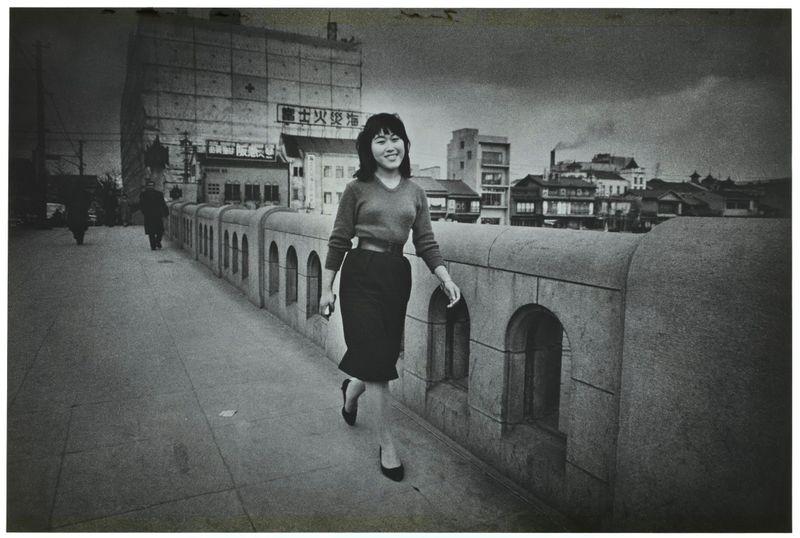 A black and white photograph depicting a woman walking along an urban bridge. She is dressed in mid-20th-century attire, with a fitted sweater paired with dark knee-length skirt and shoes. Her hair is styled straight down to her shoulders, complementing the era's fashion trends. The cityscape behind includes various buildings indicative of older architectural styles typical for Japanese cities during that period. There are faint clouds visible in the sky above, suggesting an overcast day or possible early evening ambiance as indicated by soft lighting. In the background, a building bears signage with Asian characters, which could suggest commercial establishments present at this location. The photo seems to be part of Ed van der Elsken's collection and is identified based on its provenance from the mentioned source link.