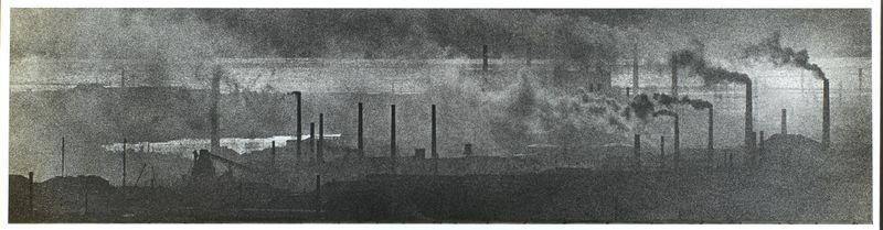 The image is a black and white panoramic photograph depicting an industrial landscape, likely from the early to mid-20th century. It shows numerous smokestacks emitting dark clouds of smoke into the sky above what appears to be factories or large industrial plants. The scene suggests heavy industry with high levels of pollution.

The photo has a grainy texture typical of older film photographs and lacks sharpness, which adds to its historical feel. There is no visible human presence in the shot; instead, the focus is on the industrial environment itself. Some structures appear silhouetted against an overcast sky or heavy smog, while others are more detailed due to lighter smoke emissions.

Given the date range provided (1959-60) and photographer Ed van der Elsken's known work within urban environments and social issues of that era, this image likely comments on industrialization's environmental impact. It could be a visual representation from Japan given the reference context stati