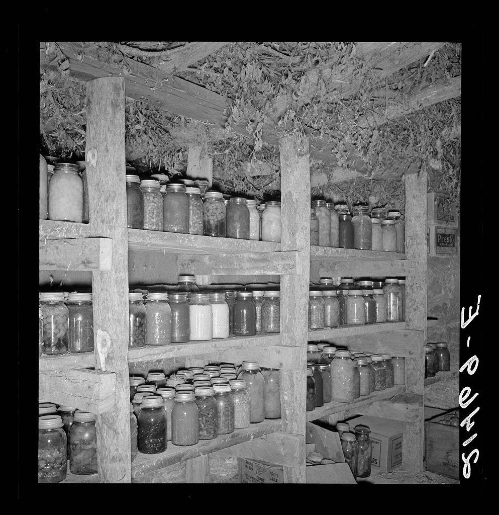 The image displays a black and white photograph of what appears to be an old-fashioned pantry or storage room, likely from the early 20th century. Numerous glass jars filled with various contents are neatly arranged on wooden shelves that extend across the space. The topmost shelf contains large mason-type jars possibly containing flour or other grain products, while smaller jars below could hold preserved foods such as pickles or jams.

The ceiling is made of exposed wood beams and rafters, suggesting a rustic construction method typical for rural homes at the time. There are dried plants hanging from one side, likely used in food preservation methods like dehydrated herbs. In the bottom right corner, there's an inscription that reads "Mrs. Wardlow 500 quarts of food," indicating Mrs. Dorothea L. Lange photographed this scene for a collection or publication.

The background is filled with more jars and possibly other storage containers on lower shelves which are not as clearly visible due to the depth-of-field used in the photograph. The overall atmosphere evokes an era where canning and preserving food was crucial, likely during times of economic hardship such as the Great Depression mentioned by Mrs. Lange herself.

The image is sourced from a website named "MrsWardlow" with additional context provided at the bottom which gives credit to Dorothea Lange for her work in  [...]